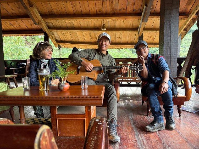 Carole sitting with two men, one playing guitar, in a wooden hut in Vietnam